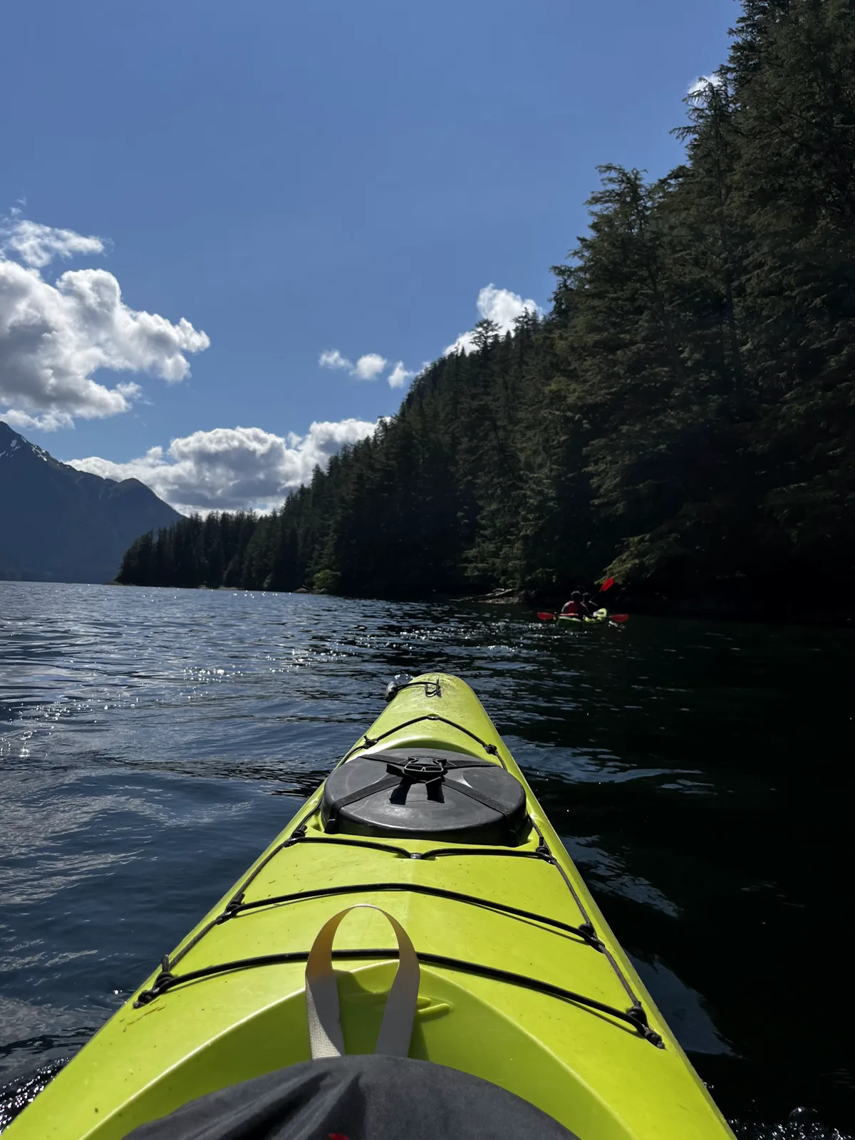 View down a kayak toward a forested shoreline and distant mountains on a sunny paddle near Sitka, Alaska