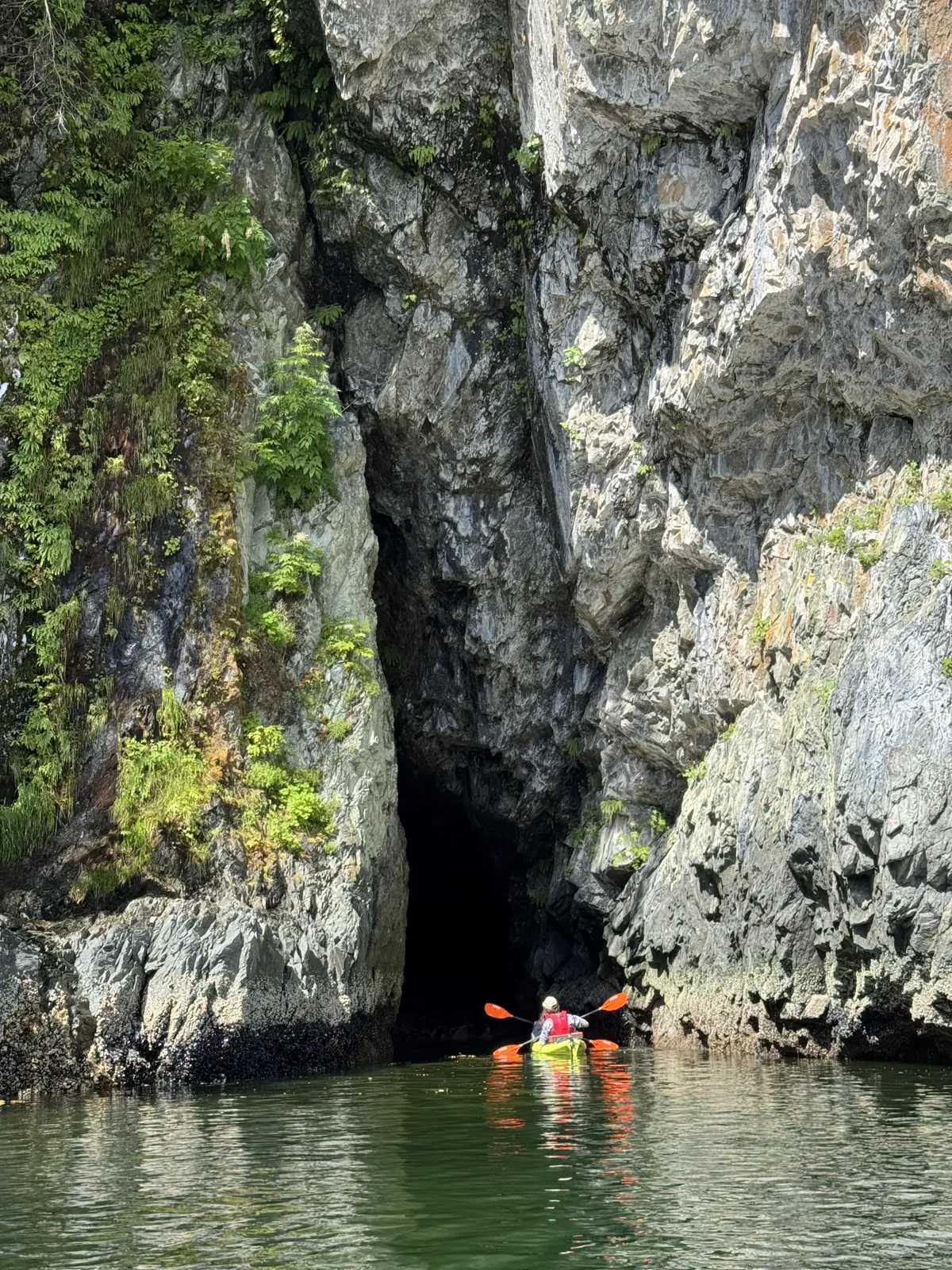Kayaker paddling toward the mouth of a sea cave in a towering rock cliff near Sitka, Alaska